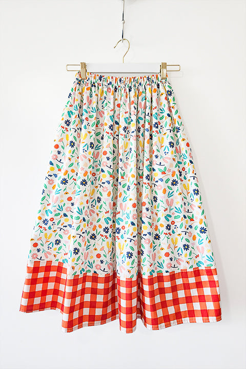 image of a colorful floral and plaid cottagecore aesthetic skirt hanging in front of a white background.  The skirt is made from 100% cotton materials and features a bright floral print paired with a red and white gingham check or plaid.