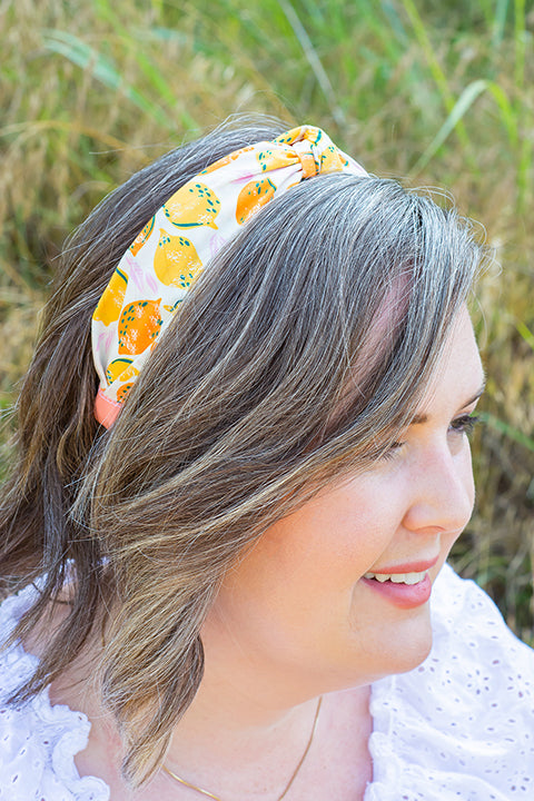 image of a female model wearing a handmade top knot headband, crafted from upcycled fabric scraps. The print on this headband is a fruit motif with retro inspired yellow lemons and oranges on a white background.