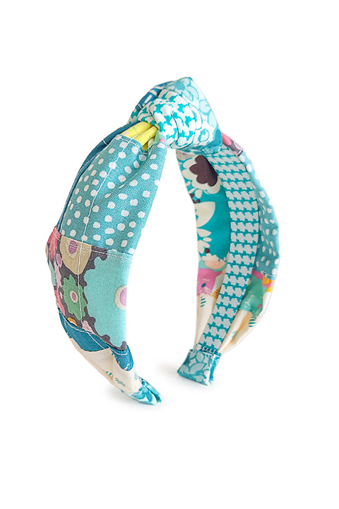 image of a handmade top knot style fabric headband.  The material on the headband is a one-of-a-kind textile created from patchworking cotton scrap fabric.  The prints are in a teal, aqua, and white color palette with florals, houndstooth, dots, cats and geometric prints