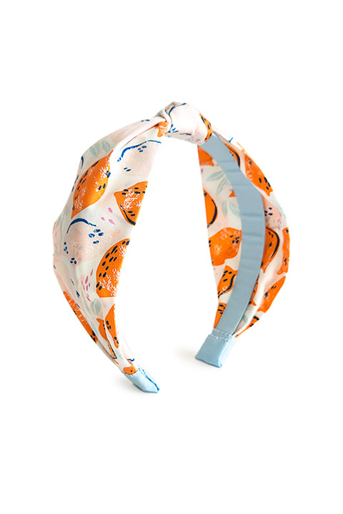 image of a top knot style handmade headband in front of a white background. this headband was sustainably made using upcycled cotton material in a lemon themed print featuring pink and orange fruit with light blue accents..