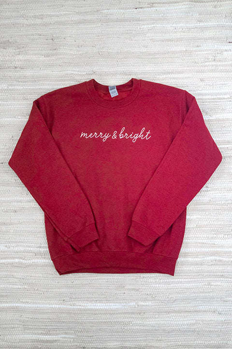 flat lay image of an antique cherry red gildan heavy blend crewneck sweatshirt screenprinted with merry & bright in a white script font