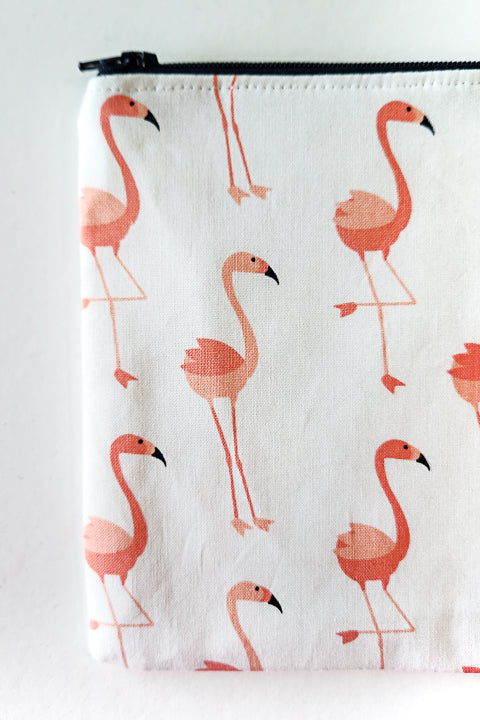 a zoomed in image of a classic zip pouch that is approximately 7.5" w x 6" tall.  The zip pouch is flat and made with a black zipper and 100% cotton material.  The print for this bag features cute pink flamingos on a white background.