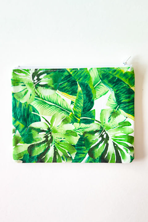 image of a flat 100% cotton handmade zip pouch. The bag is 7.5" wide by 6" tall.  It features a white zipper and a green tropical leaves print.