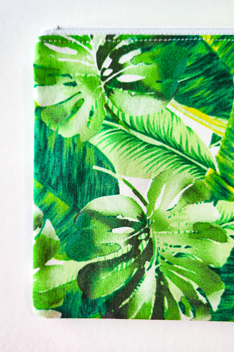 a zoomed in detailed image of a flat 100% cotton zip pouch. The bag is 7.5" wide by 6" tall.  It features a white zipper and a green tropical leaves print.