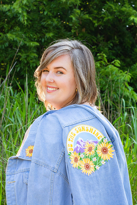 smiling white female model looking over shoulder while holding a light blue denim jacket that has a large embroidered patch sewn onto the back.  the back patch says be the sunshine arched over mountains and sunflowers. the jacket also has a small yellow sunflower patch sewn onto the front left lapel of the jacket.