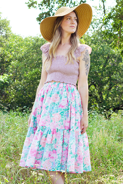 image of a size small female model standing in a meadow wearing a sunhat, smocked sleeveless top and a handmade vintage textile skirt.  The midi skirt features a pastel pink, green and blue cabbage rose floral design and has an elastic waist and ruffled hem.