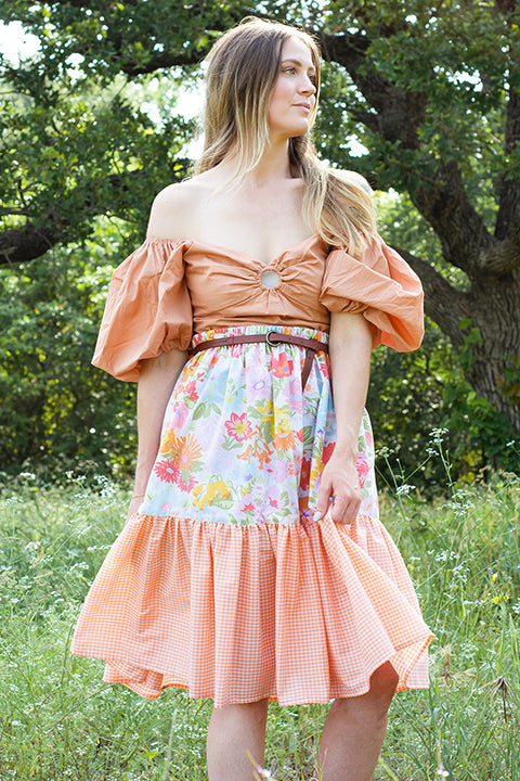 image of a size small female model standing in a treelined field of flowers wearing a romantic off the shoulder puff sleeve top paired with a one-of-a-kind ruffled midi skirt made from upcycled vintage materials.  The skirt has a bright, colorful retro floral print paired with a small peach and white gingham check for the ruffle.