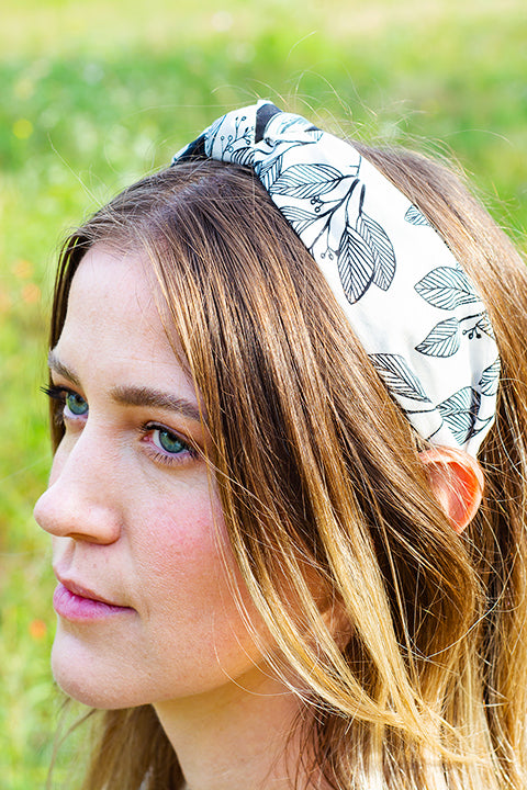 image of a female model wearing a top knot headband, handmade using upcycled cotton fabric scraps.  This headband is split color, with one print on one half of the headband and a coordinating print on the other.  This side of the headband is black linear leaves printed on white.