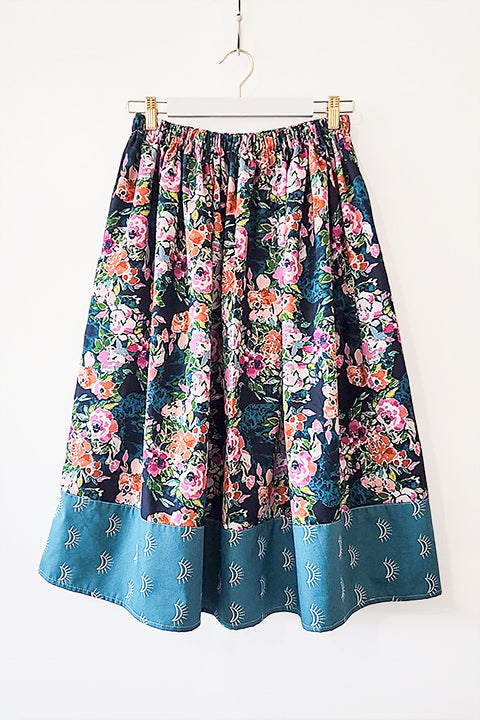 image of a cottagecore aesthetic midi length skirt that features a bold, bright and romantic floral print paired with a bottom border in a sunmoon boho design.  The colors in the fabrics are dark teals with accents of pink, white, coral and green.
