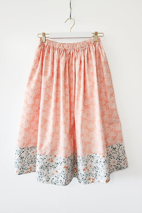 image of a midi length cotton skirt hanging in front of a white wall.  The skirt has a cottagecore vibe and features floral themed materials.  The main material features white ferns on a pastel blush pink background.  The border material at the hem features watercolor impressionist inspired florals in mint, sage green and pale pink,