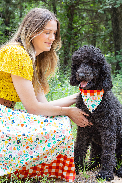 image of a dog mom bending over and petting her black poodle dog.  The woman is wearing a skirt that coordinates with her dog's reversible bandana.  The main print for the skirt and bandana is a bright, colorful floral on a white background.  The accent print on the skirt and reverse side of the bandana is a red and white gingham picnic plaid.