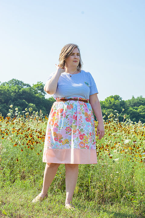 image of a plus size female model standing in front of a field of wildflowers wearing a casual cottagecore inspired outfit.  The model is wearing a secondhand tee paired with a one-of-a-kind skirt made from vintage materials.  The skirt has a vibrant 60s floral print paired with a peach and white mini gingham check.