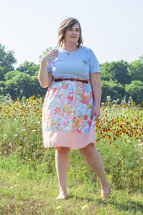 image of a plus size female model standing in front of a field of wildflowers wearing a casual cottagecore inspired outfit. The model is wearing a secondhand tee paired with a one-of-a-kind skirt made from vintage materials. The skirt has a vibrant 60s floral print paired with a peach and white mini gingham check.