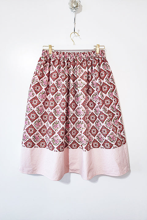 image a of a midi length skirt hanging in front of a white wall.  The skirt is sustainably made from 2 one of a kind vintage textiles.  The main skirt fabric is a red, pink and green diamond pattern floral print.  The trim of the skirt is a mini gingham check in light pink and white.