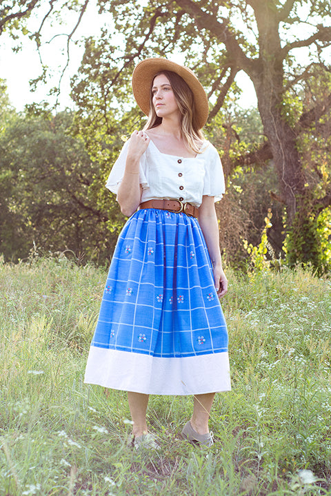 image a of a white, blonde female model standing in a tree lined meadow with the sun softly backlighting her as she looks down. The model is a wearing a cottagecore aesthetic outfit: a white flowy linen button down top, paired with a high waisted midi length skirt and wide belt. The skirt is one-of-a-kind made from vintage cotton materials and is mainly a blue cotton windowpane plaid paired with a white daisy lace