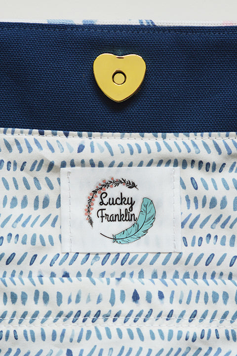 a close up image showing the details of the lining of the tote bag.  the interior shows a brass or gold colored heart shape magnetic snap closure, a logo label that say lucky franklin, the facing material which is dark blue canvas, and the lining fabric which is a lightweight white cotton printed with light blue watercolor slash lines..