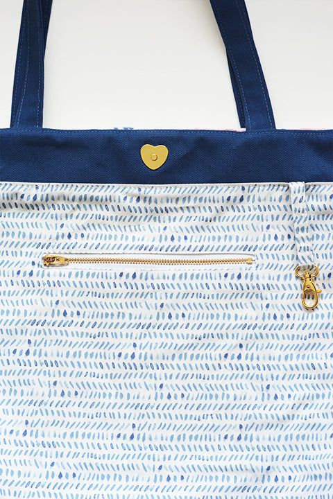 a detailed image of the inside of the tote bag that shows an interior pocket with a metal zip closure, a hook hanging from a piece of fabric that is for holding keys, the gold brass heart shaped magnetic snap closure, the dark blue denim handles and facing, and the lining fabric which is white cotton printed with light blue watercolor lines.