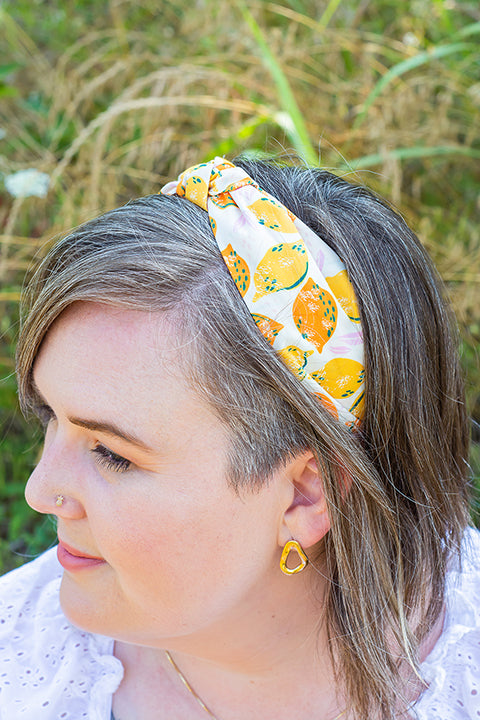 image of a female model wearing a handmade top knot headband, crafted from upcycled fabric scraps.  The print on this headband is a fruit motif with retro inspired yellow lemons and oranges on a white background.