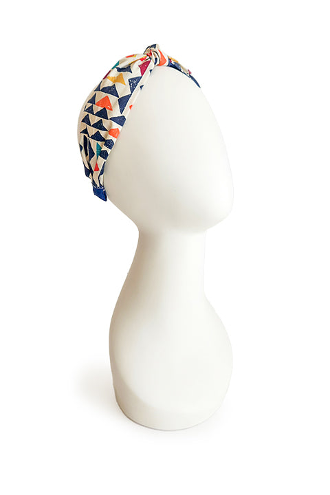 image of a sustainable and upcycled top knot style headband shown on a white head display in front of a white background. This headband was handmade using fabric scraps.  The 100% cotton material on this headband features a geometric triangle print in a boho color palette of mustard yellow, navy blue, orange, teal, and cardinal red.