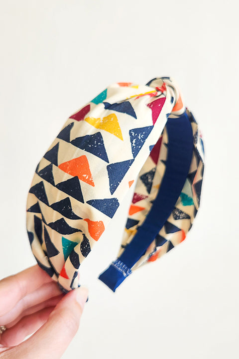 image of a hand holding a sustainable and upcycled top knot style headband in front of a white background. This headband was handmade using fabric scraps.  The 100% cotton material on this headband features a geometric triangle print in a boho color palette of mustard yellow, navy blue, orange, teal, and cardinal red.