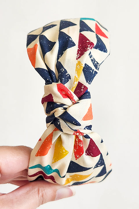 image of a hand holding a sustainable and upcycled top knot style headband in front of a white background. This headband was handmade using fabric scraps.  The 100% cotton material on this headband features a geometric triangle print in a boho color palette of mustard yellow, navy blue, orange, teal, and cardinal red.