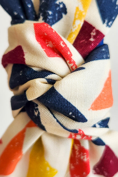 a super zoomed in view of the twisted top knot of a sustainable and upcycled top knot style headband in front of a white background. This headband was handmade using fabric scraps.  The 100% cotton material on this headband features a geometric triangle print in a boho color palette of mustard yellow, navy blue, orange, teal, and cardinal red.
