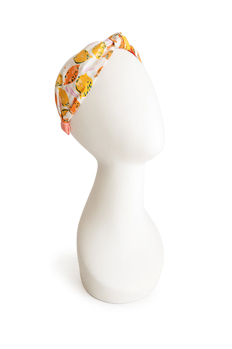 image of a top knot style handmade headband shown on a white head display in front of a white background. this headband was sustainably made using upcycled cotton material in a lemon themed print featuring orange and yellow fruit.