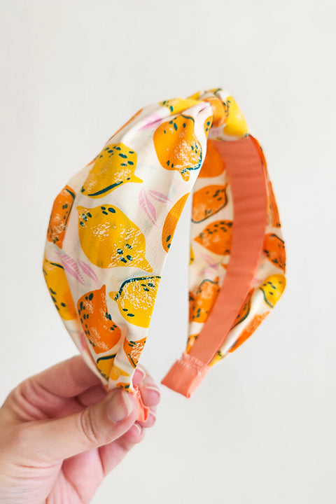 image of hand holding a top knot style handmade headband in front of a white background. this headband was sustainably made using upcycled cotton material in a lemon themed print featuring orange and yellow fruit.