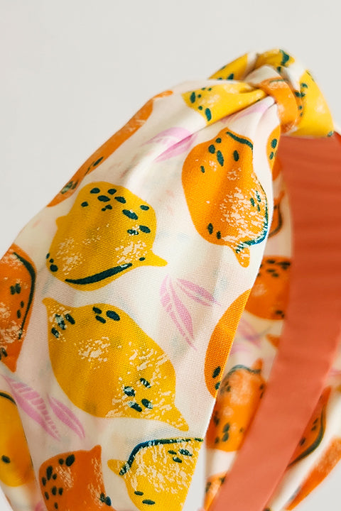 a zoomed in detailed image of a top knot style handmade headband in front of a white background. this headband was sustainably made using upcycled cotton material in a lemon themed print featuring orange and yellow fruit.