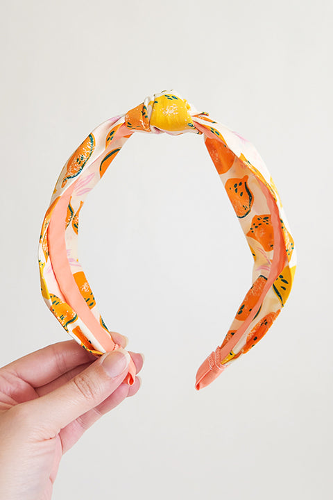 image of a hand holding a top knot style handmade headband in front of a white background. this headband was sustainably made using upcycled cotton material in a lemon themed print featuring orange and yellow fruit.
