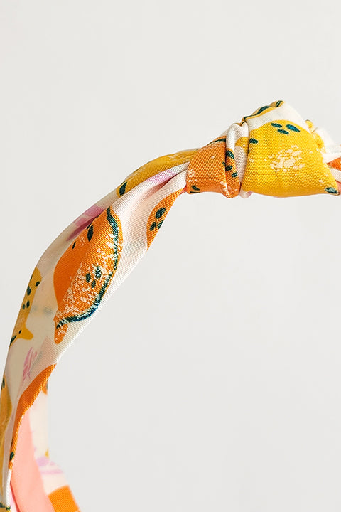 a detailed image of a top knot style handmade headband in front of a white background. this headband was sustainably made using upcycled cotton material in a lemon themed print featuring orange and yellow fruit.