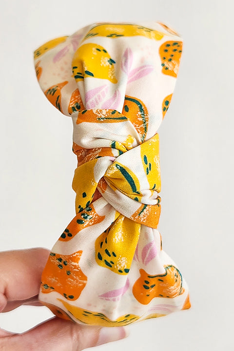 an image of a hand holding a top knot style handmade headband in front of a white background. this headband was sustainably made using upcycled cotton material in a lemon themed print featuring orange and yellow fruit.