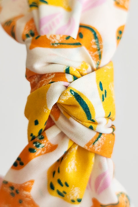 a zoomed in detailed image of the top of top knot style handmade headband in front of a white background. this headband was sustainably made using upcycled cotton material in a lemon themed print featuring orange and yellow fruit.