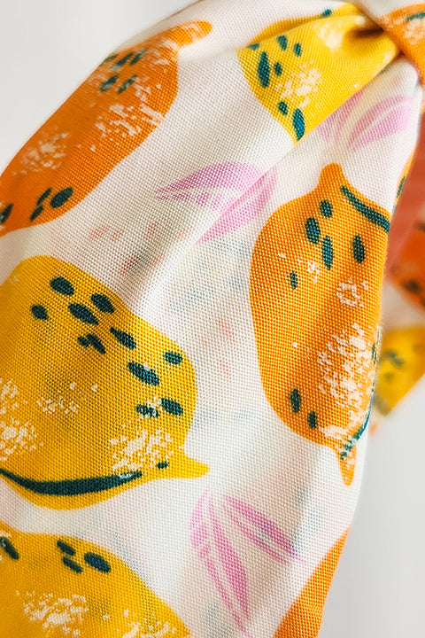 a detailed view of the fabric of a top knot style handmade headband in front of a white background. this headband was sustainably made using upcycled cotton material in a lemon themed print featuring orange and yellow fruit.