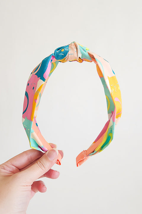 image of a hand holding a  top knot style headband shown in front of a white background. the headband is sustainably handmade from cotton fabric scraps in a color cat themed print.  the colors in the cat print are magenta, teal, aqua, yellow and orange.