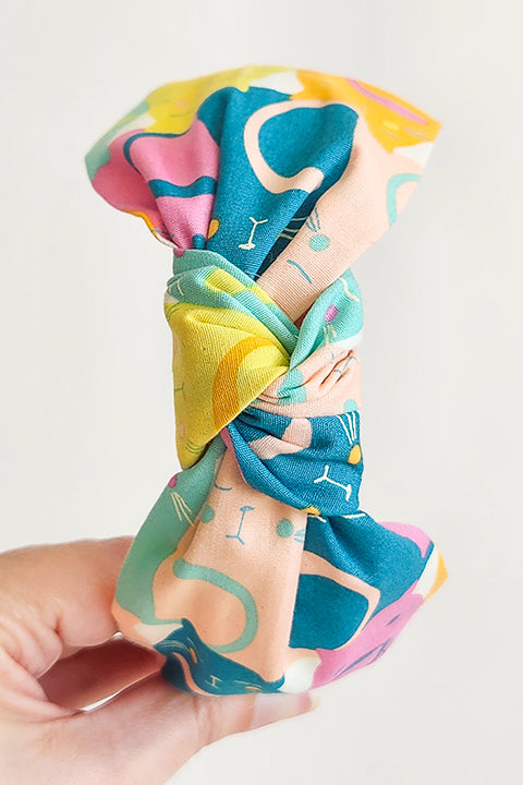 image of a hand holding a top knot style headband shown in front of a white background. the headband is sustainably handmade from cotton fabric scraps in a color cat themed print.  the colors in the cat print are magenta, teal, aqua, yellow and orange.