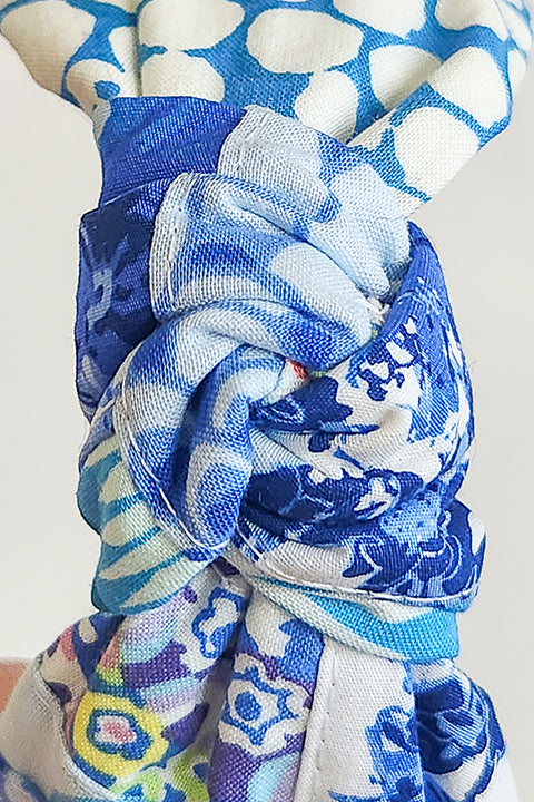 a close up, very zoomed in image of the twisted top knot a handmade and sustainable top knot style headband in front of a white background.  The cotton material on the headband is a unique patchworked textile created from scrap fabrics in shades of blue with white.  The prints feature florals, dots, geometric shapes and buttons.