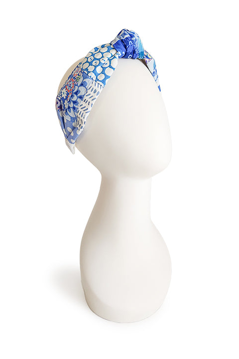 an image of a handmade and sustainable top knot style headband shown on a white head display in front of a white background.  The cotton material on the headband is a unique patchworked textile created from scrap fabrics in shades of blue with white.  The prints feature florals, dots, geometric shapes and buttons.