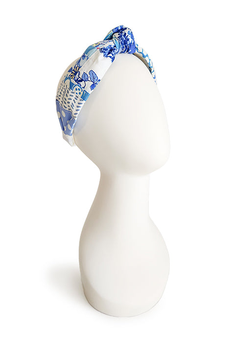 an image of a handmade and sustainable top knot style headband shown on a white head display in front of a white background.  The cotton material on the headband is a unique patchworked textile created from scrap fabrics in shades of blue with white.  The prints feature florals, dots, geometric shapes and buttons.