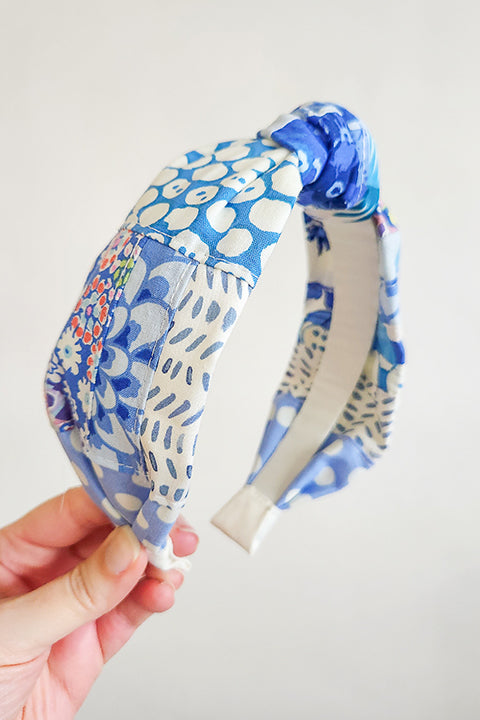 an image of a hand holding a handmade and sustainable top knot style headband in front of a white background.  The cotton material on the headband is a unique patchworked textile created from scrap fabrics in shades of blue with white.  The prints feature florals, dots, geometric shapes and buttons.