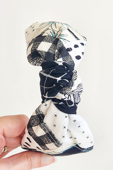 image of a hand holding a handstitched top knot style fabric headband in front of a white background.  The material on the headband is a one-of-a-kind textile created from patchworking cotton scrap fabric.  The prints are in a black and white color palette with florals, checks, stripes, dots and triangles. headband is shown from the top.