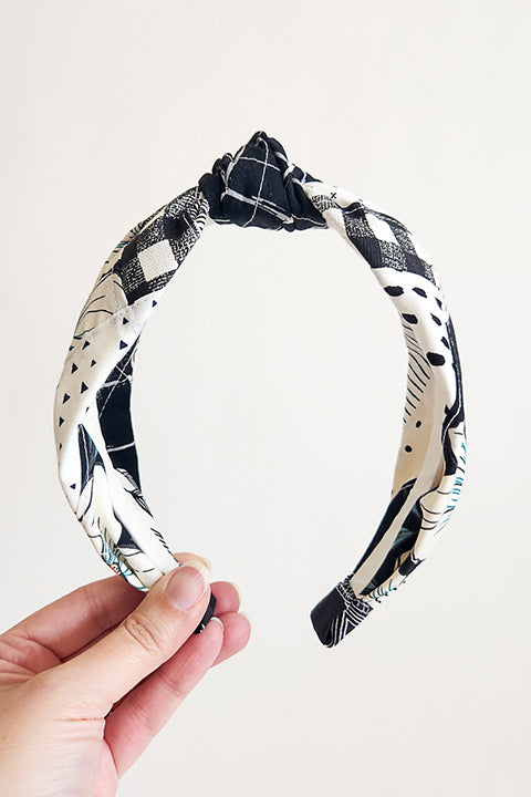 image of a hand holding a handstitched top knot style fabric headband in front of a white background.  The material on the headband is a one-of-a-kind textile created from patchworking cotton scrap fabric.  The prints are in a black and white color palette with florals, checks, stripes, dots and triangles