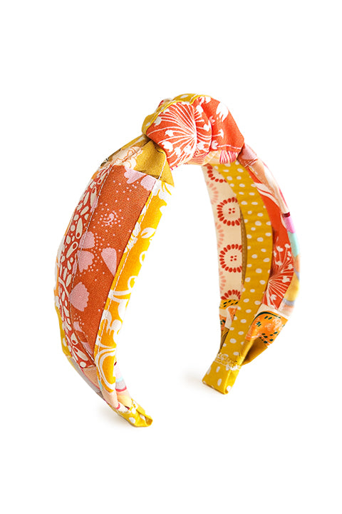 image of a hand stitched top knot style fabric headband.  The material on the headband is a one-of-a-kind textile created from patchworking cotton scrap fabric.  The prints are in an orange, pink and yellow color palette with florals, dots and geometric prints.