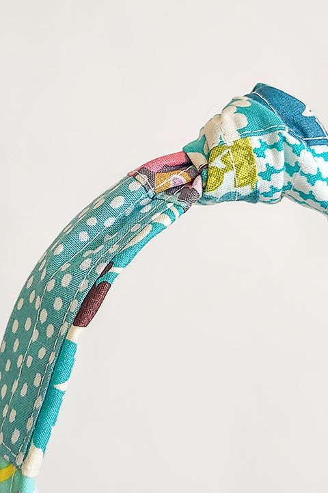 detailed zoomed in image a handmade top knot style fabric headband in front of a white background. The material on the headband is a one-of-a-kind textile created from patchworking cotton scrap fabric. The prints are in a teal, aqua, and white color palette with florals, houndstooth, dots, cats and geometric prints