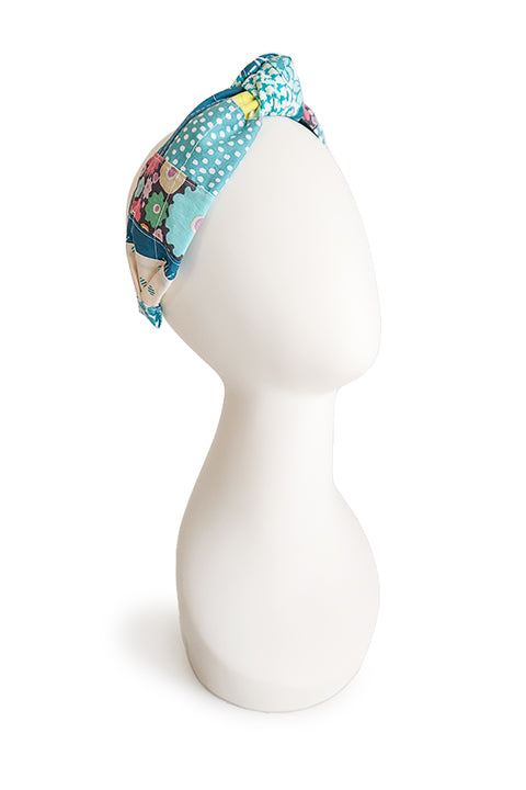 image of a handmade top knot style fabric headband shown on a white colored display head in front of a white background.  The material on the headband is a one-of-a-kind textile created from patchworking cotton scrap fabric.  The prints are in a teal, aqua, and white color palette with florals, houndstooth, dots, cats and geometric prints