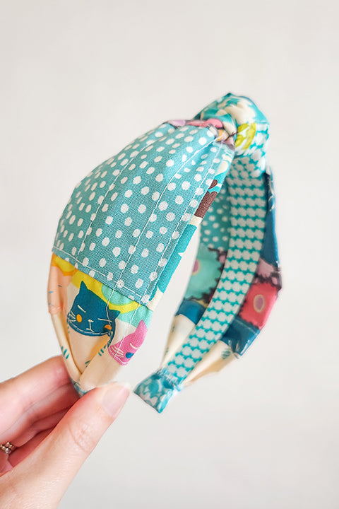 image of a hand holding a handmade top knot style fabric headband in front of a white background. The material on the headband is a one-of-a-kind textile created from patchworking cotton scrap fabric. The prints are in a teal, aqua, and white color palette with florals, houndstooth, dots, cats and geometric prints