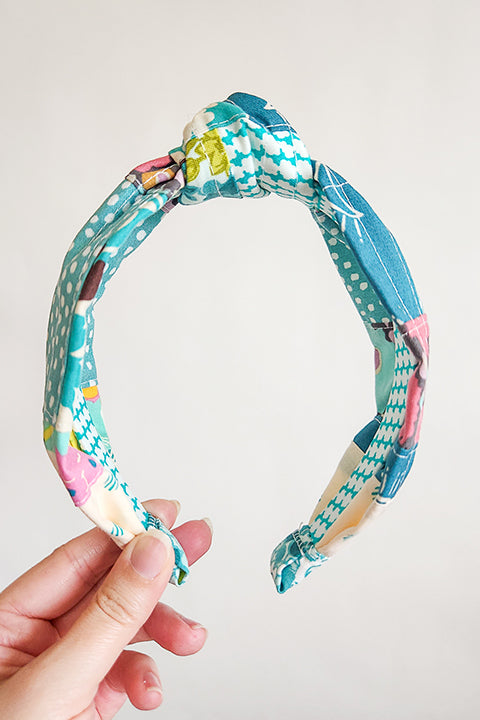 image of a hand holding a handmade top knot style fabric headband in front of a white background. The material on the headband is a one-of-a-kind textile created from patchworking cotton scrap fabric. The prints are in a teal, aqua, and white color palette with florals, houndstooth, dots, cats and geometric prints