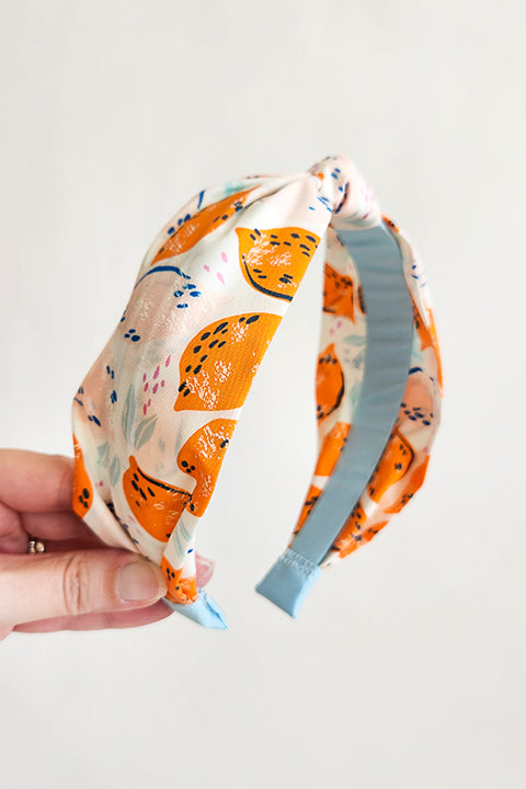image of a hand holding a top knot style handmade headband in front of a white background. this headband was sustainably made using upcycled cotton material in a lemon themed print featuring pink and orange fruit with light blue accents..