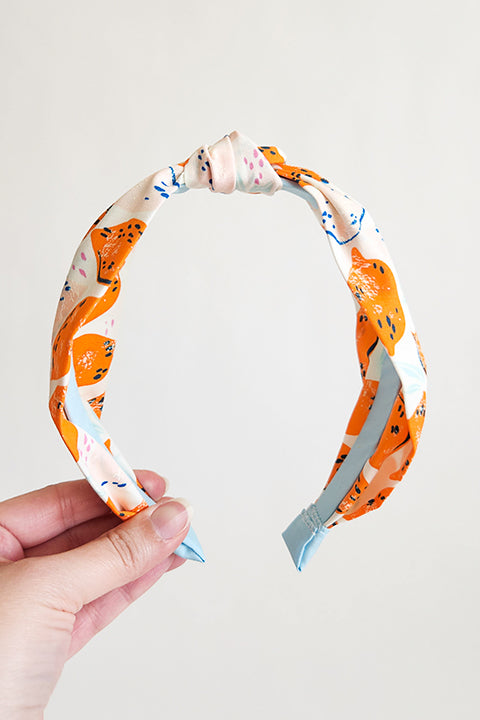 image of a hand holding a top knot style handmade headband in front of a white background. this headband was sustainably made using upcycled cotton material in a lemon themed print featuring pink and orange fruit with light blue accents..