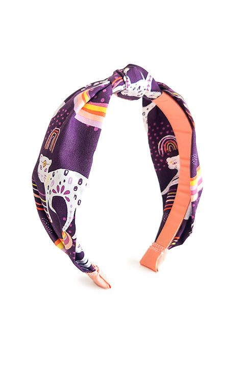 image of a top knot styled handmade headband in front of a white background.  Sustainably made from 100% cotton fabric scraps the print features boho white jaguars and yellow, pink and coral rainbows on a dark purple background.